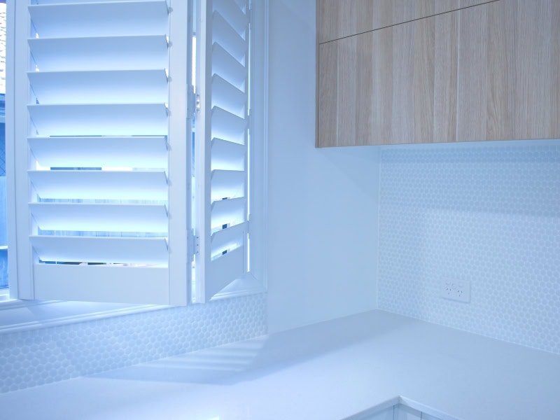 White plantation shutters in kitchen beside wood cabinets and white benchtop.