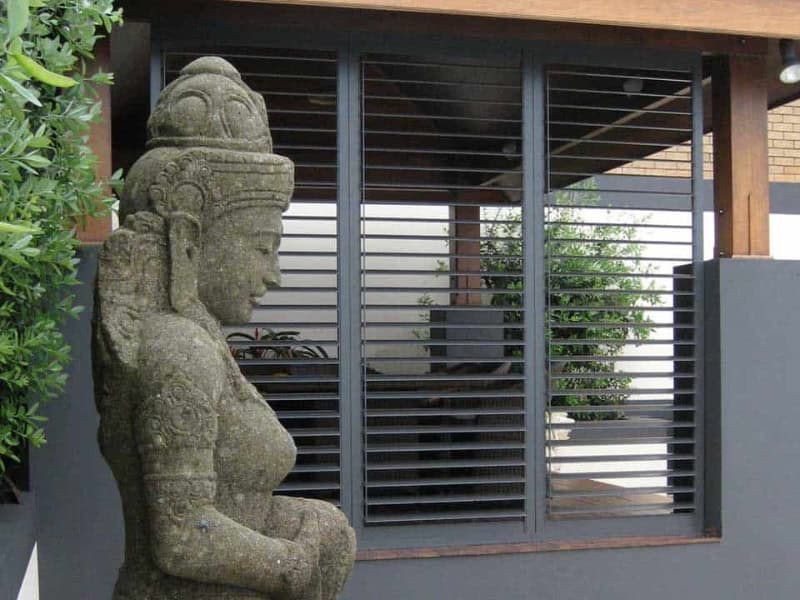 A modern outdoor living space with sleek black plantation shutters behind a decorative stone statue and timber pergola.