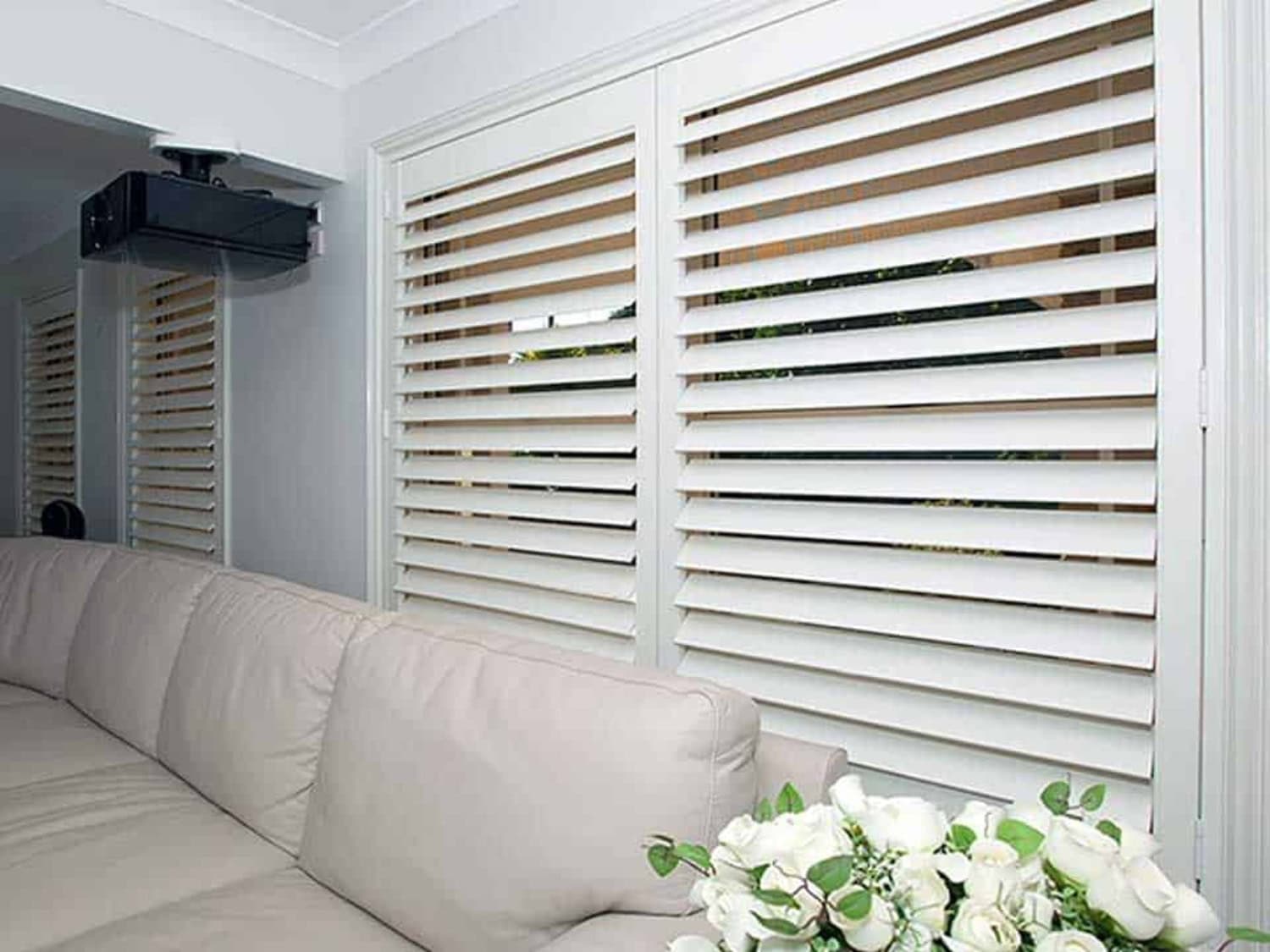 White plantation shutters for big windows behind a lounge area with a sofa and flowers.