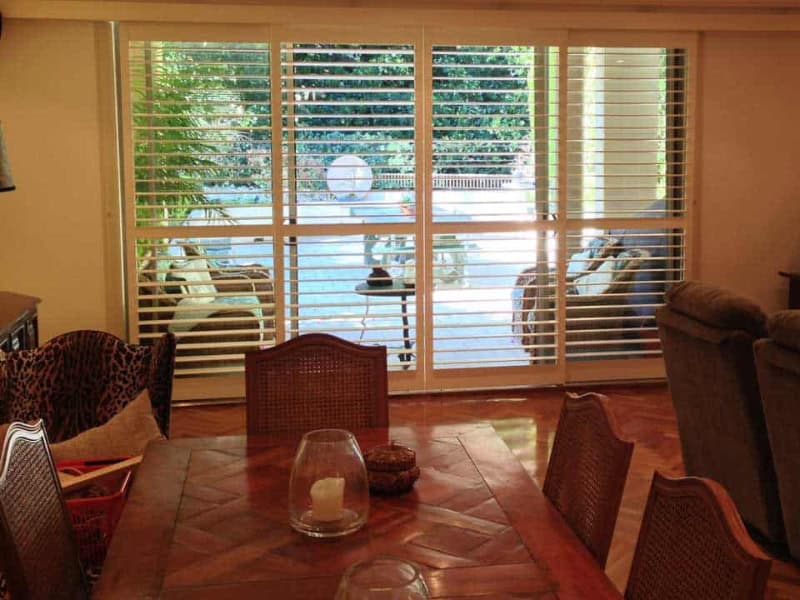 Large sliding doors fitted with white outdoor shutters for decks overlooking a patio with seating and greenery.