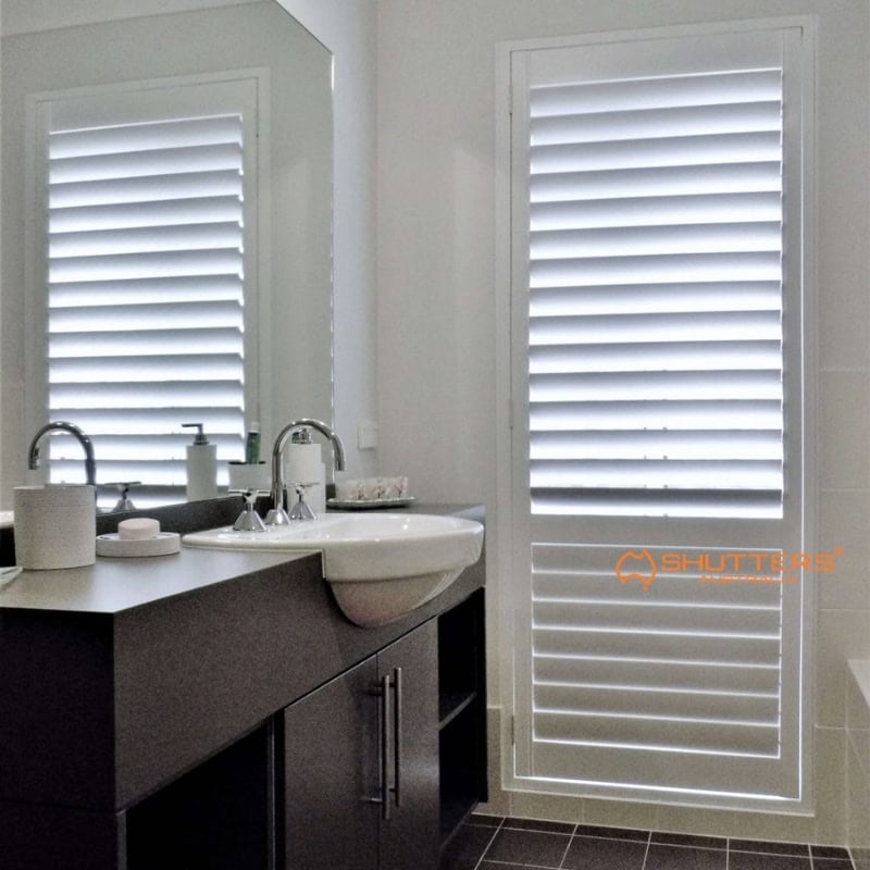 Interior plantation shutters on tall bathroom window beside modern vanity and mirror, white louvres filtering natural light.