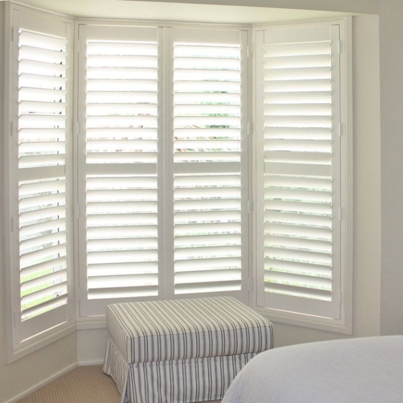 Plantatio shutters on bright bay window with white louvres and cushioned bench seat in a modern bedroom.