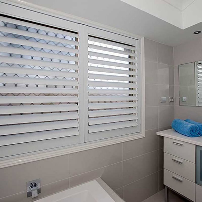 A bathroom window featuring installed white plantation Shutters, providing privacy over textured glass, with a view of a bathtub.
